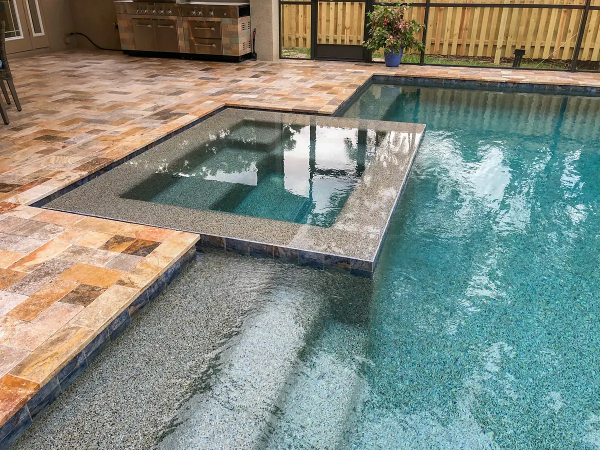 Square spa integrated into a turquoise swimming pool with stone paving inside a screened patio.