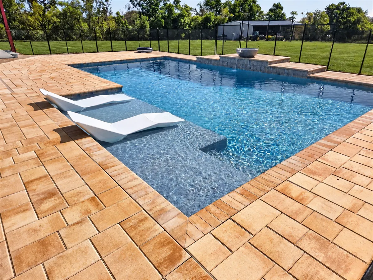 Sunlit swimming pool with two white loungers on a shallow tanning ledge, surrounded by stone pavers.